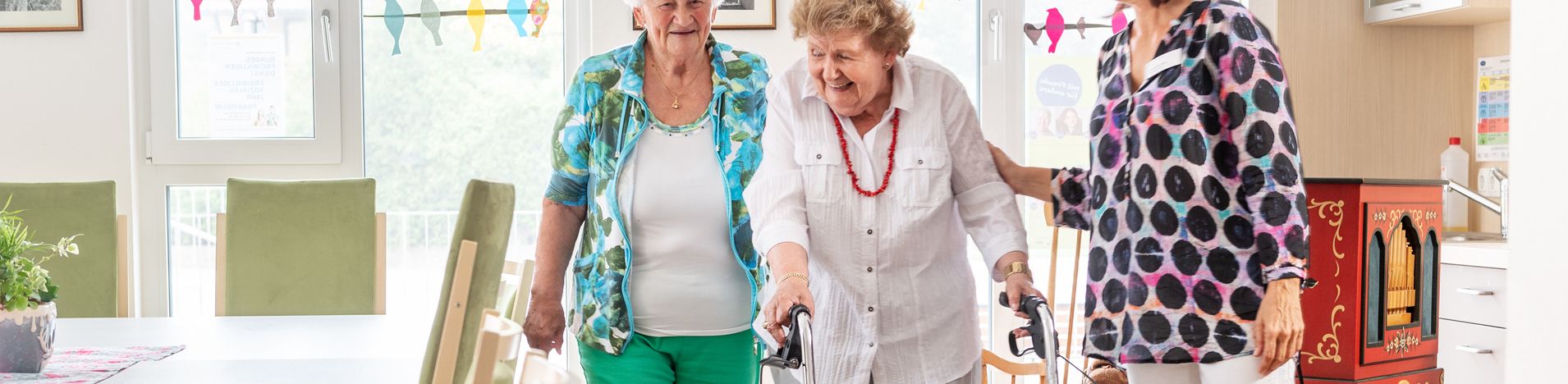 Ältere Frau mit Rollator wird von zwei Frauen begleitet