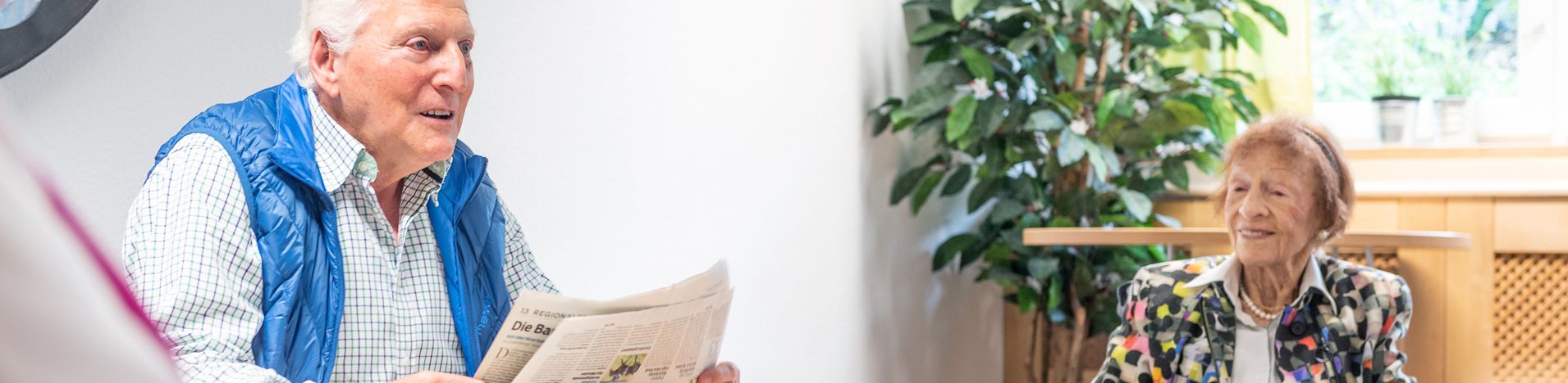 Älterer Mann liest Zeitung und ältere Frau im Rollstuhl hört zu